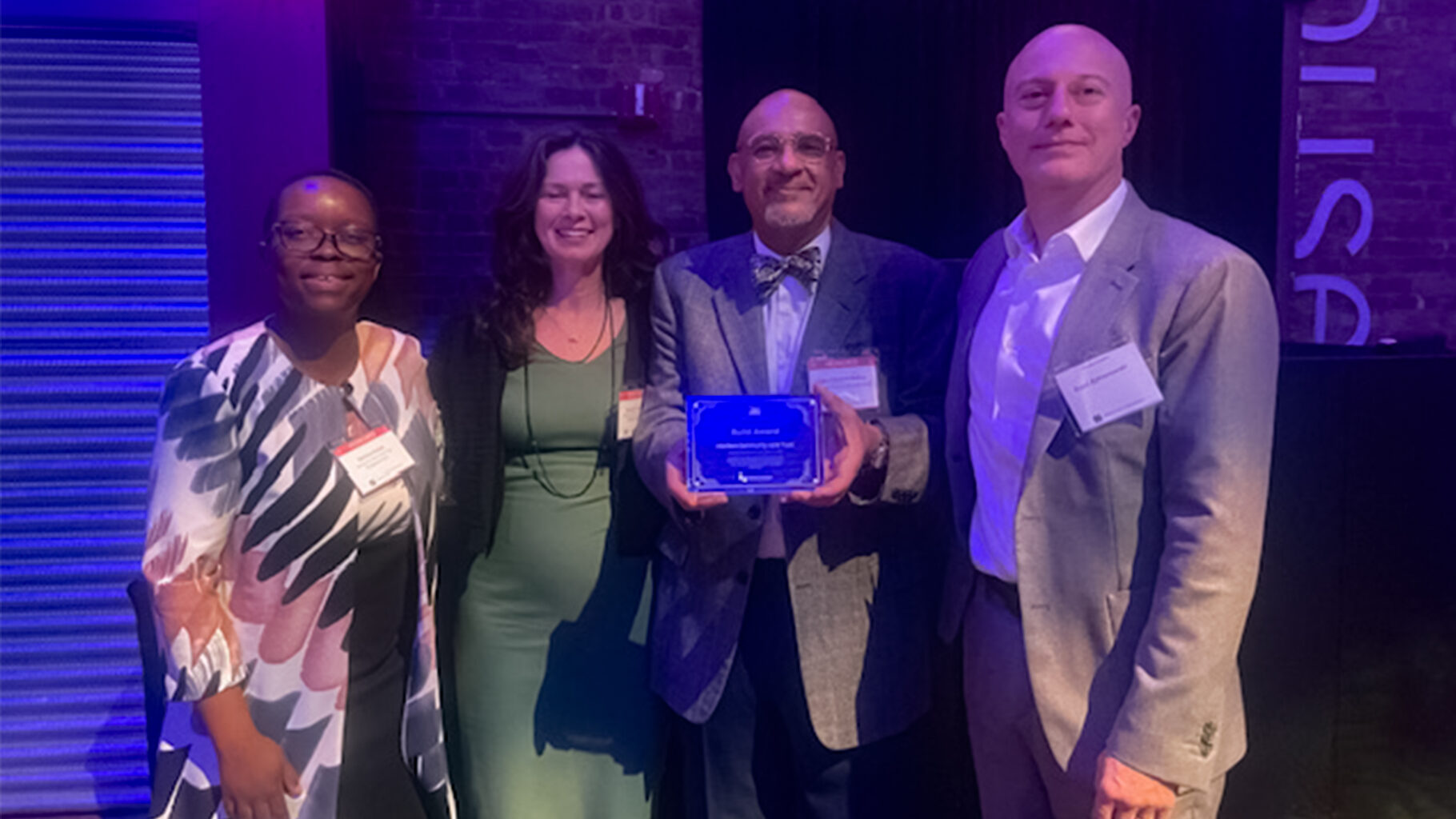 From left to right: Salima Etoka, Center for NYC Neighborhoods Chief of Staff; Christie Peale, Center CEO and Interboro Community Land Trust Board Chair; John Edward Dallas, Director, Interboro; and Scott Kohanowski, General Counsel for the Center. John Edward holds the Build Award plaque. All are smiling and dressed in formal attire under purple event lighting.